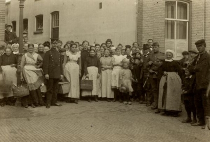 Op de foto wachtende mensen onder toezicht van een agent en een militair voor het distributielokaal aan de gasfabriek op de Havenweg; fotograaf Gebroeders Janssen.